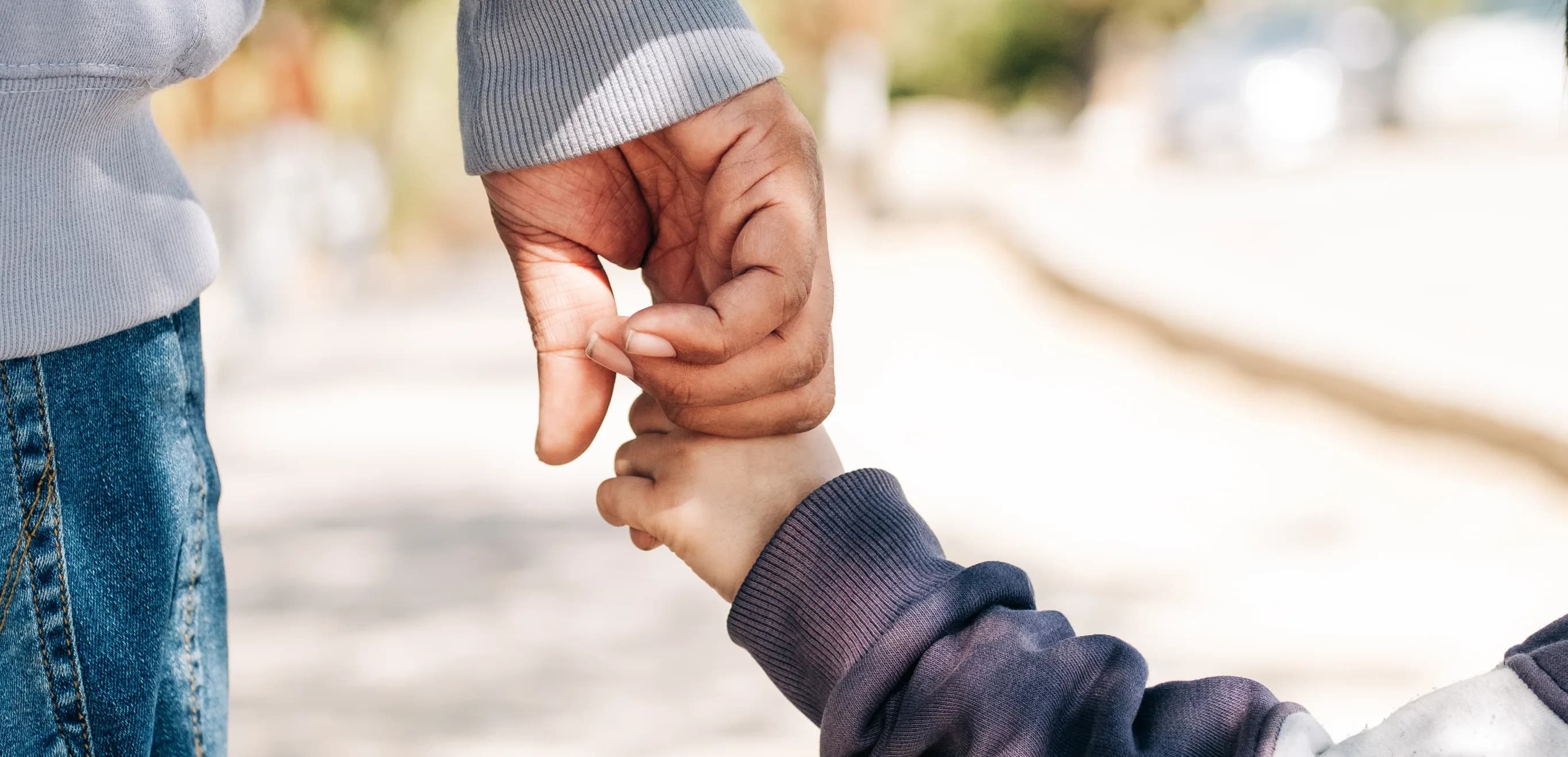 An adult's hand gently holding a young child's hand outdoors, both in warm knitwear, on a softly blurred street