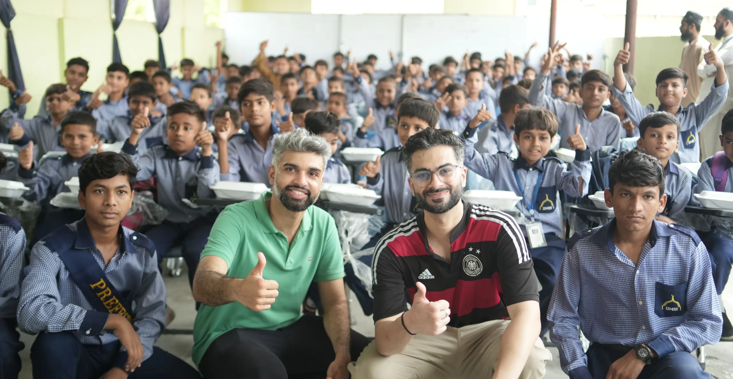Two Shukar Hai founders give a thumbs-up in front of a school hall full of uniformed boys with meal boxes on their desks