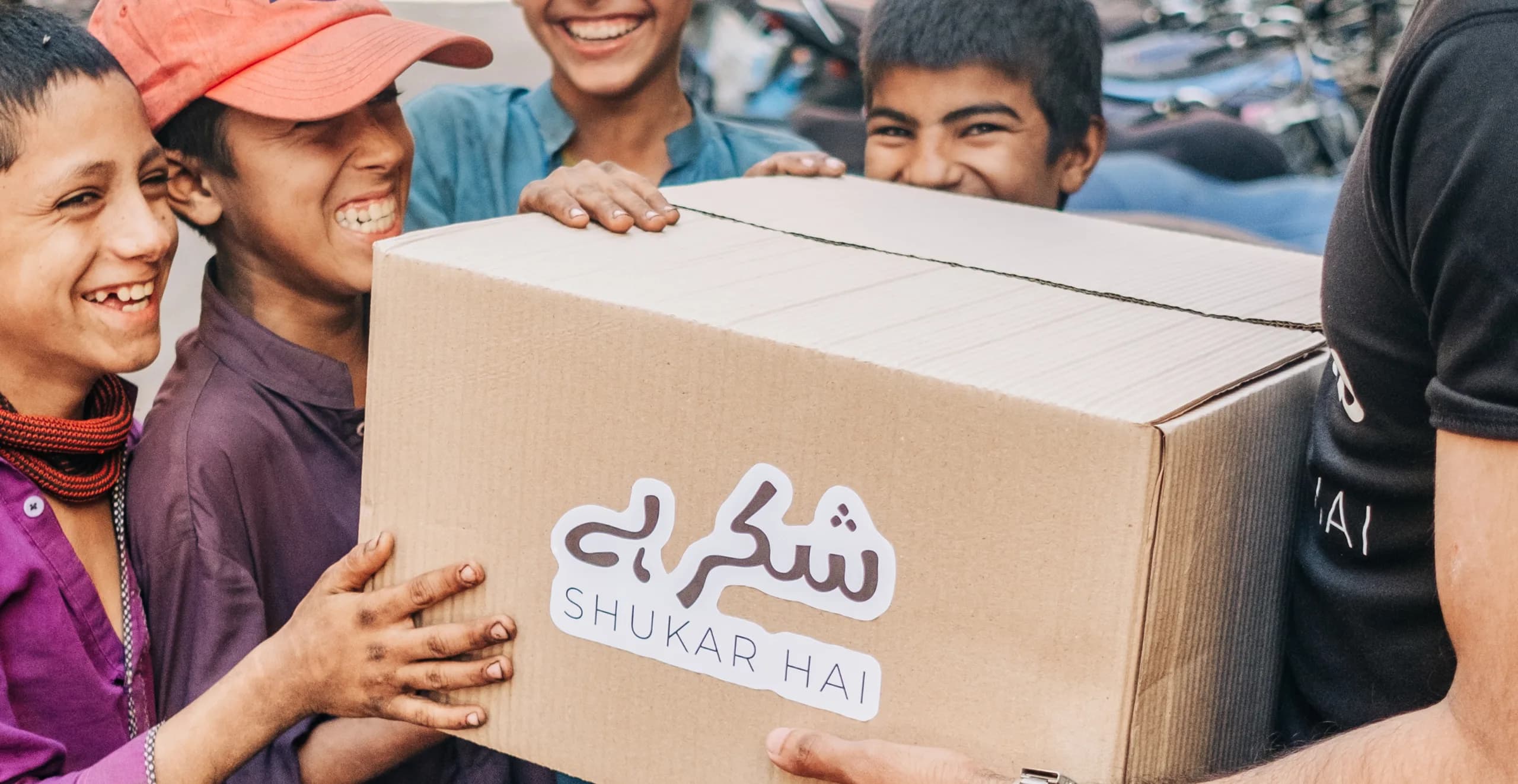 Four laughing boys help carry a large Shukar Hai delivery box down a street